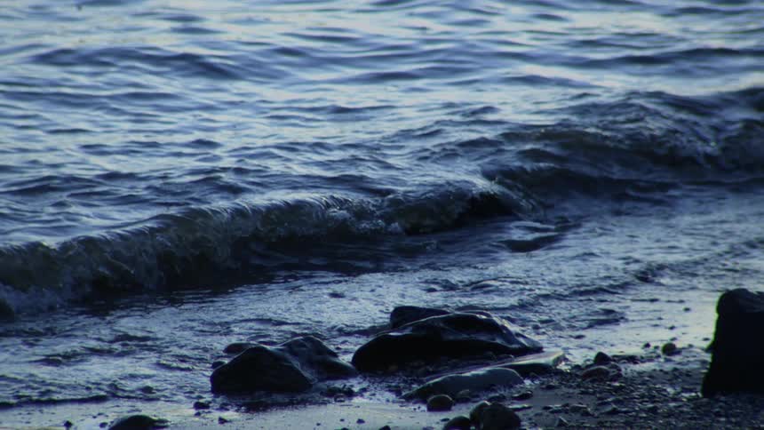 Slow motion shot of gentle dark blue waves lapping against a rocky shore