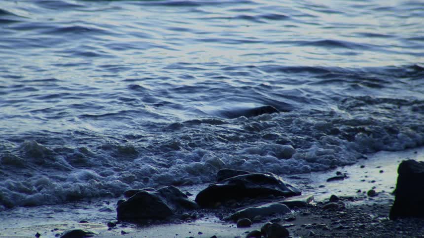 Dark blue waves lap against a rocky shore in moody slow motion