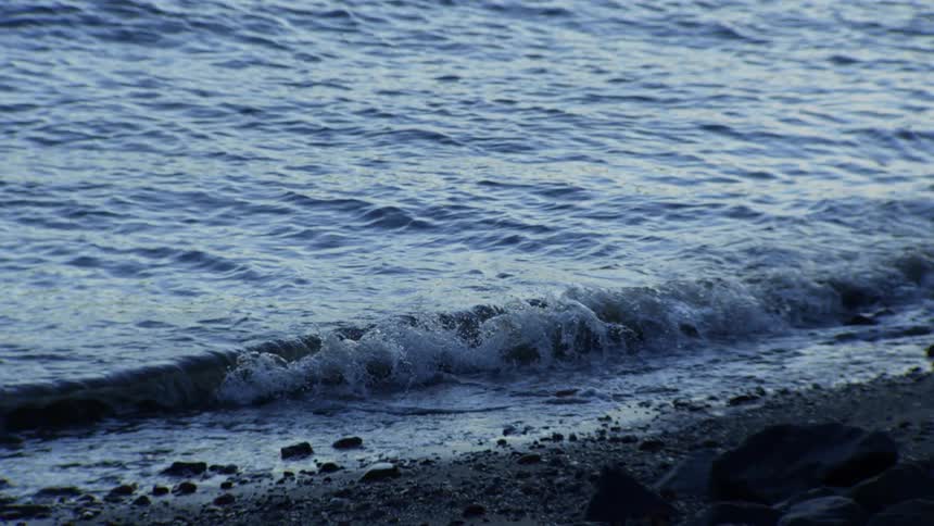 A serene, super slow-motion close-up of a small wave gently breaking on a rocky beach at dusk