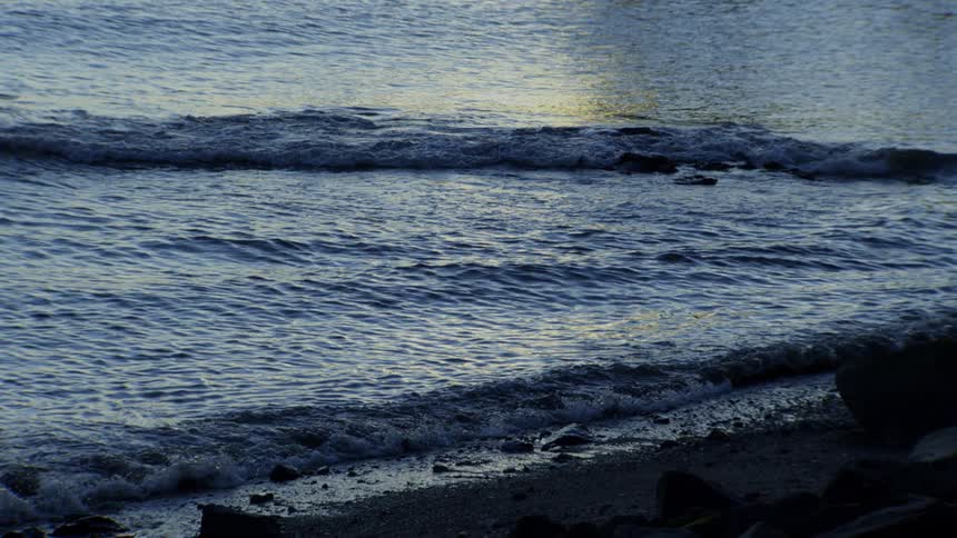 Calm blue waves lap against a rocky shoreline in mesmerizing slow motion at twilight