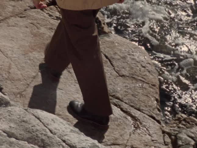 A person's legs in brown pants and black shoes are shown as they carefully step over large rocks next to a sparkling ocean