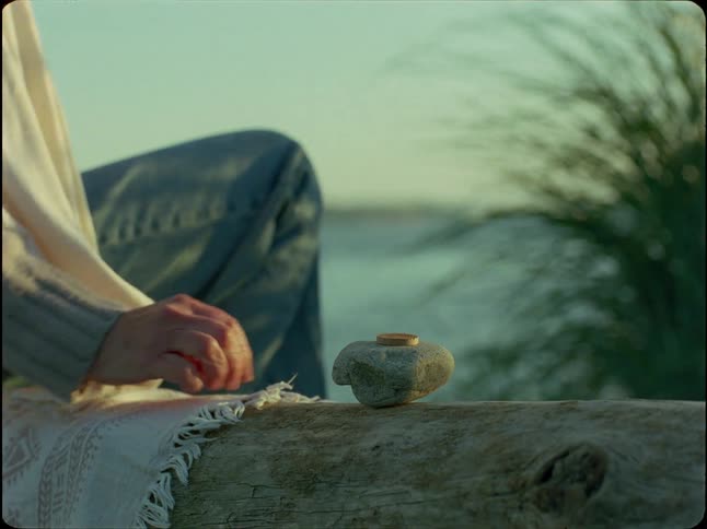 A person's hand carefully places a small stone on another rock on a driftwood log by the ocean