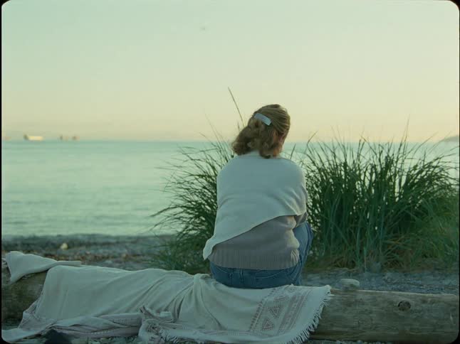 A woman with her back to the camera sits on a driftwood log, peacefully watching the ocean at dusk