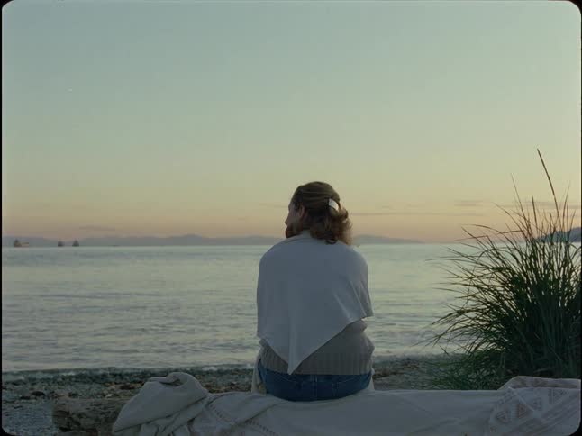 A solitary figure sits on a rocky beach, peacefully watching the calm ocean and distant ships during a serene sunset