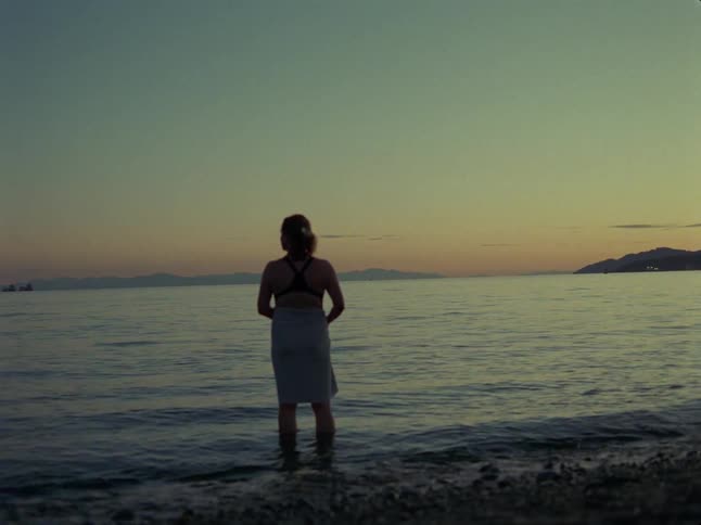 A woman wrapped in a towel stands in the shallow ocean water at sunset, peacefully gazing at the horizon