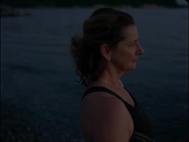 A contemplative woman in a swimsuit stands by the ocean at dusk before the camera pans to a wide, serene view of the water and distant ships