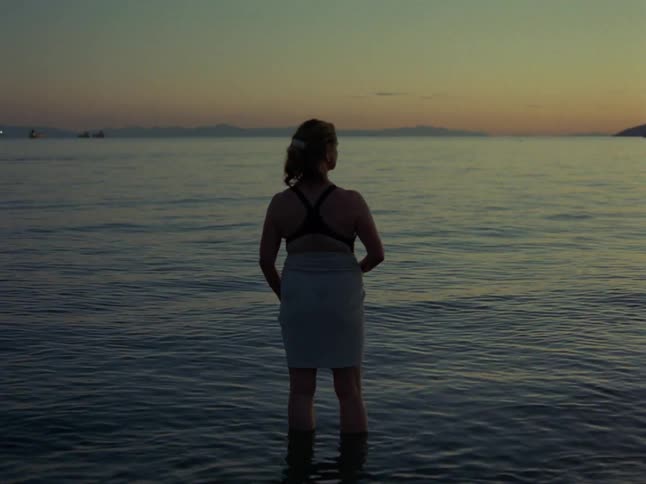 A contemplative woman with a towel wrapped around her waist stands in the calm ocean at dusk, looking out at the horizon