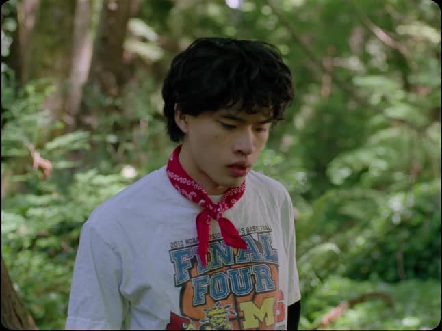 A sad young Asian man wearing a red bandana wipes a tear from his eye while standing alone in a lush green forest