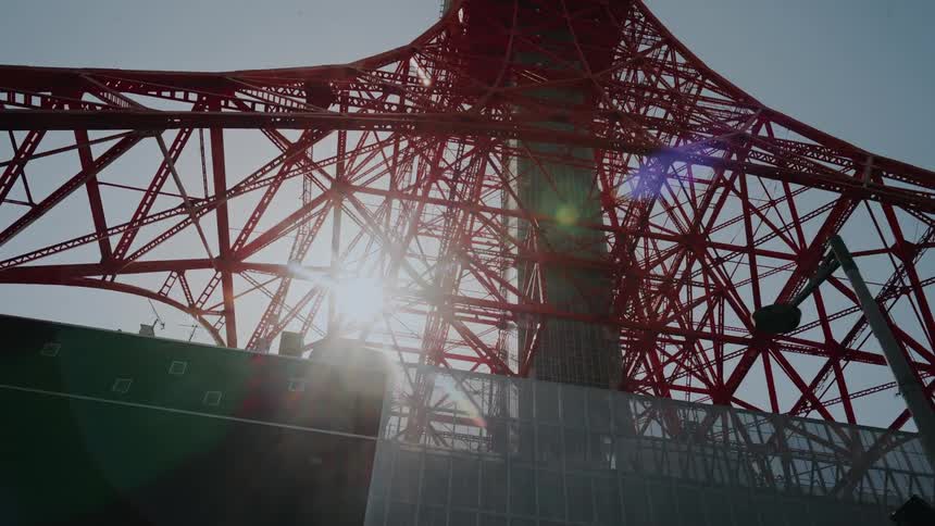 An upward-looking tracking shot of the majestic Tokyo Tower with the sun shining brightly through its red steel frame
