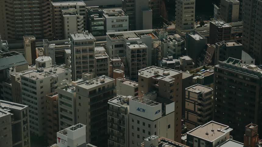 A high-angle static shot shows the dense architecture and rooftops of a bustling modern city during the day
