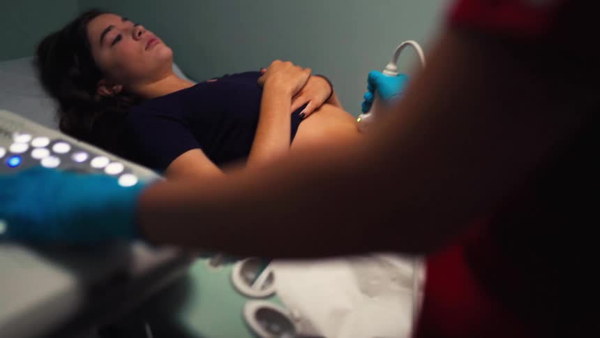 A doctor performs an abdominal ultrasound on a young woman, with the focus shifting to the machine's controls