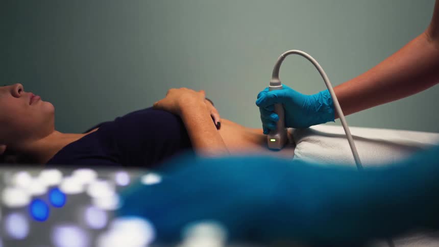 A doctor performs an abdominal ultrasound on a young female patient in a clinic, with the machine's keyboard blurred in the foreground