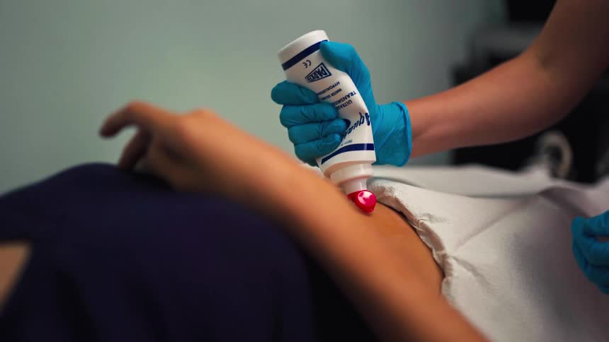 A medical professional in blue gloves applies ultrasound gel to a patient's abdomen before an examination