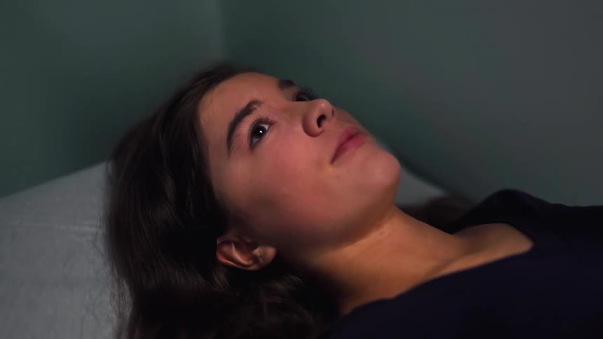 Close-up of a pensive young woman lying down in a dimly lit room, staring upwards