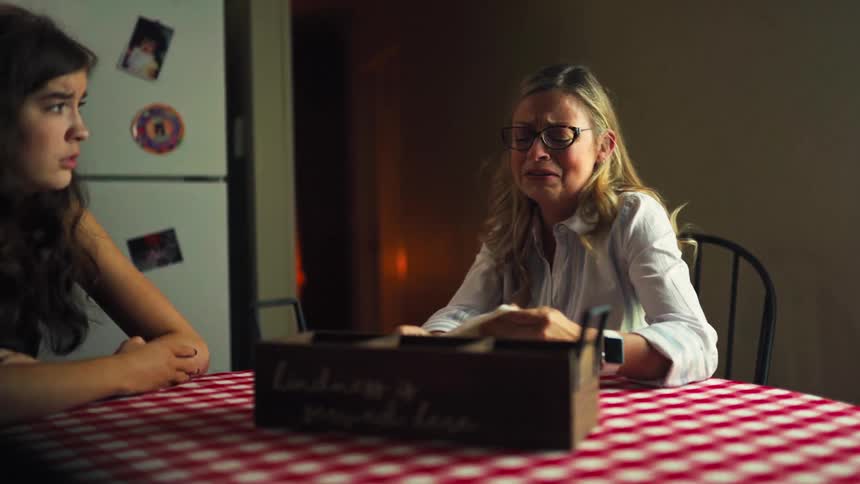 A distraught mother cries at the kitchen table while her concerned teenage daughter looks on