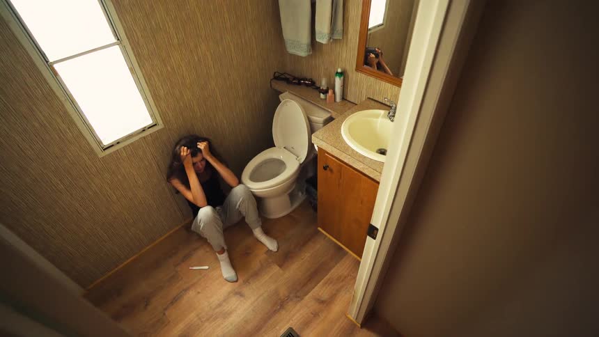 A high angle shot captures a distraught young woman sitting on the bathroom floor in despair after seeing the results of a pregnancy test