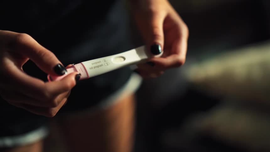 A close-up shot of a woman's hands holding a positive pregnancy test in a dimly lit room