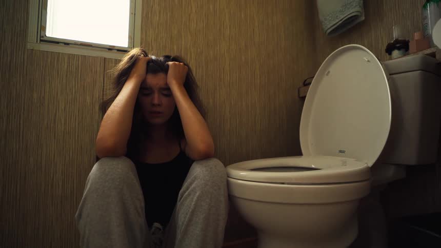 A troubled and sick young woman sits in despair on the bathroom floor next to the toilet