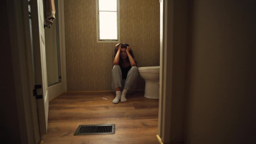 A slow pull-back shot reveals an anxious young woman sitting alone on the bathroom floor after seeing the results of her pregnancy test