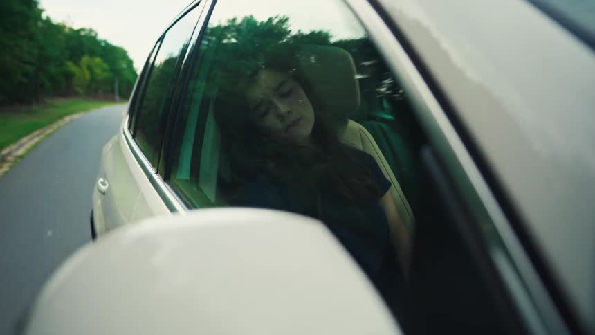 A tired young woman sleeps in the passenger seat of a moving car, with the countryside scenery reflected on the window