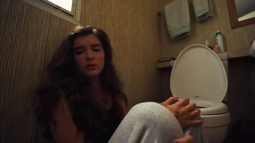 A depressed young woman with a pained expression sits on the floor of a dimly lit bathroom