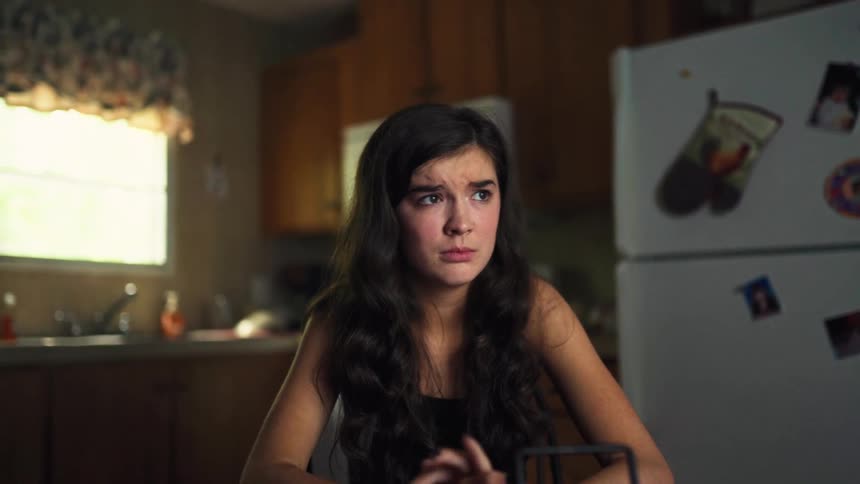 A distressed teenage girl in a kitchen shows increasing sadness and fear while looking at something on her phone
