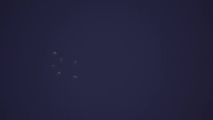 A group of white balloons are released and float away into the dark blue night sky until they disappear from view