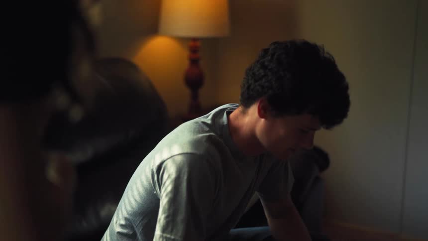An emotional young man has a heartfelt conversation, his face shifting from tears to a hopeful smile in a dimly lit room