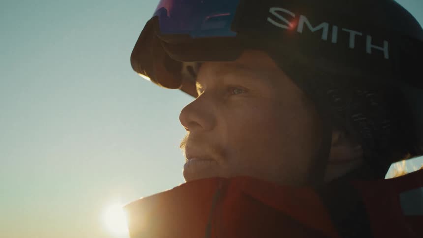 A close-up of a snowboarder's face, wearing a helmet and looking out at the horizon during a golden sunset