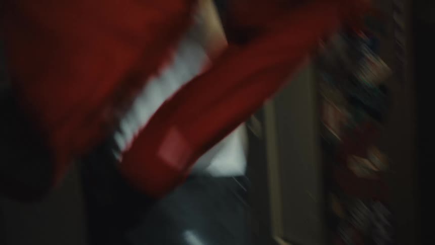 A man in a beanie gets dressed in a dark ski lodge locker room, putting on a red jacket with a Swiss cross