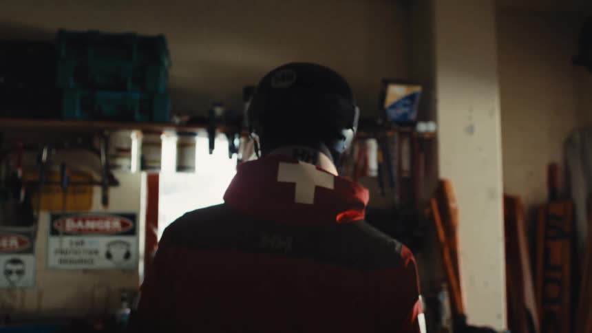 A ski patroller in full gear walks through a dimly lit workshop, viewed from behind