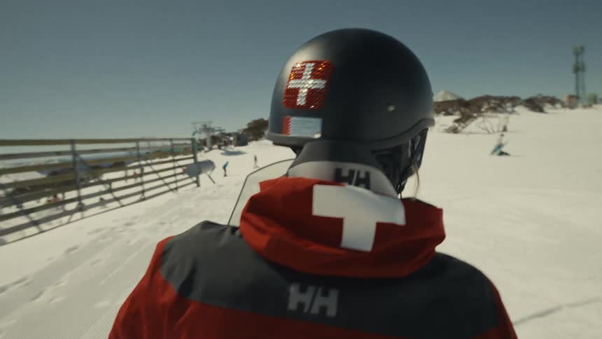 A follow shot of a ski patrol officer riding a snowmobile up a snowy mountain at a ski resort on a sunny day