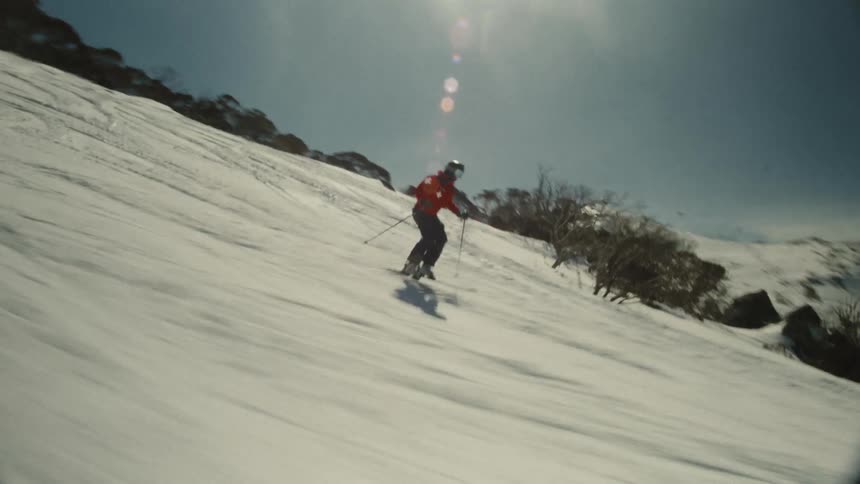 A dynamic tracking shot follows a skilled skier in a red jacket as they swiftly descend a sun-drenched, snow-covered mountain