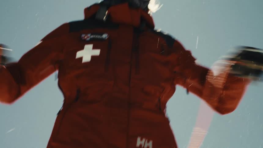 A smiling female ski patroller in full gear throws an orange rescue rope in slow motion against a bright, sunny sky
