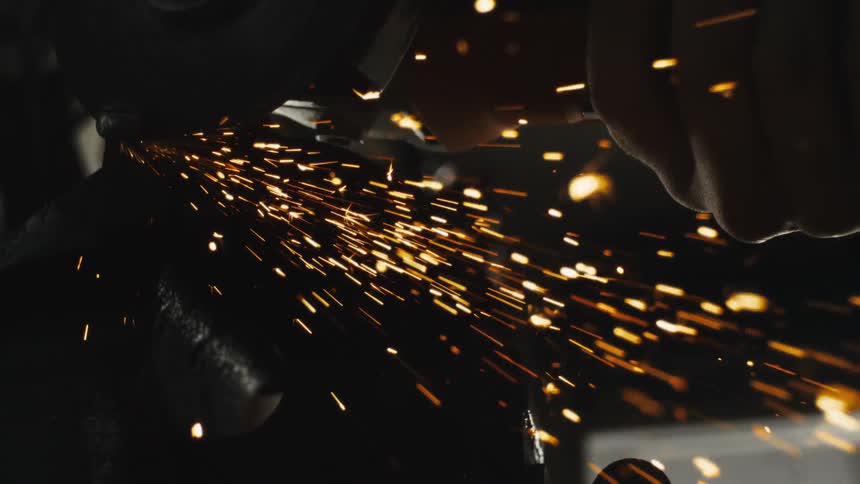 A dramatic slow-motion close-up of a metal grinder sending a shower of bright orange sparks into the darkness