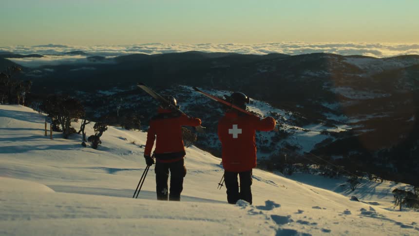 From behind, two members of the ski patrol carry their skis and look out over a stunning mountain vista at golden hour