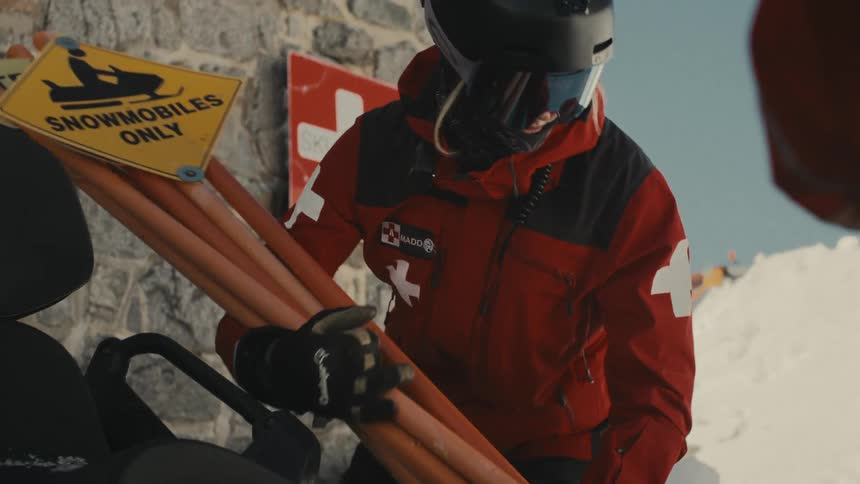 A ski patrol member in a red uniform and helmet carries trail markers next to a snowmobile at a ski resort