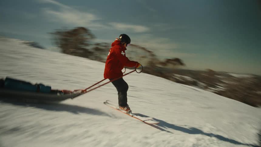 A ski patroller in a red jacket skillfully skis down a snowy mountain while pulling a rescue sled on a bright, sunny day