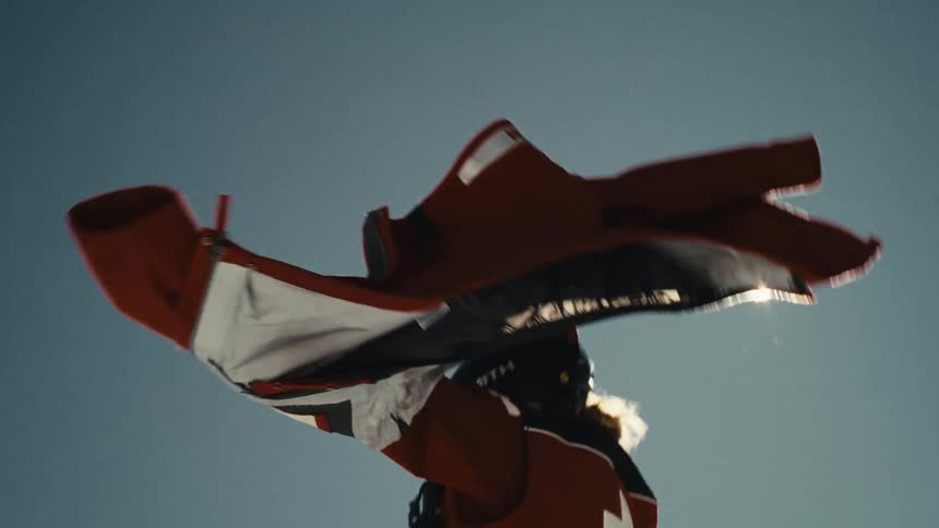 A ski patroller holds a red winter jacket aloft in the wind, letting it fly in the sun in cinematic slow motion