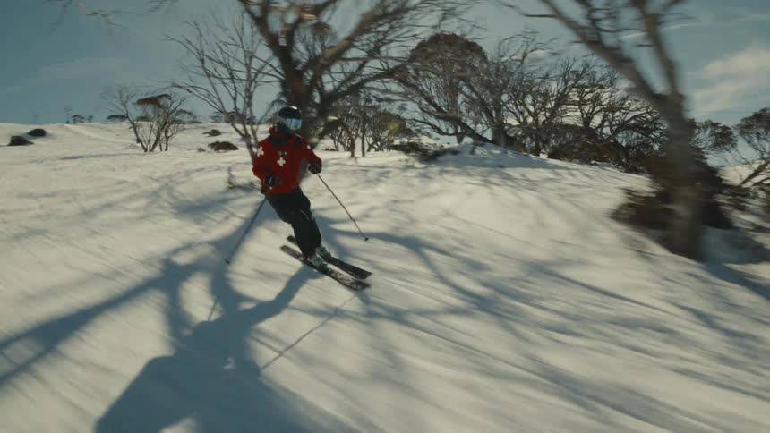A ski patroller in a red jacket skis down a sunny, snow-covered slope past bare trees at a mountain resort