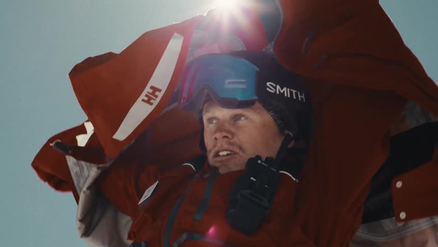 Low-angle slow-motion shot of a male skier putting on his red jacket with a bright sun flare in the background