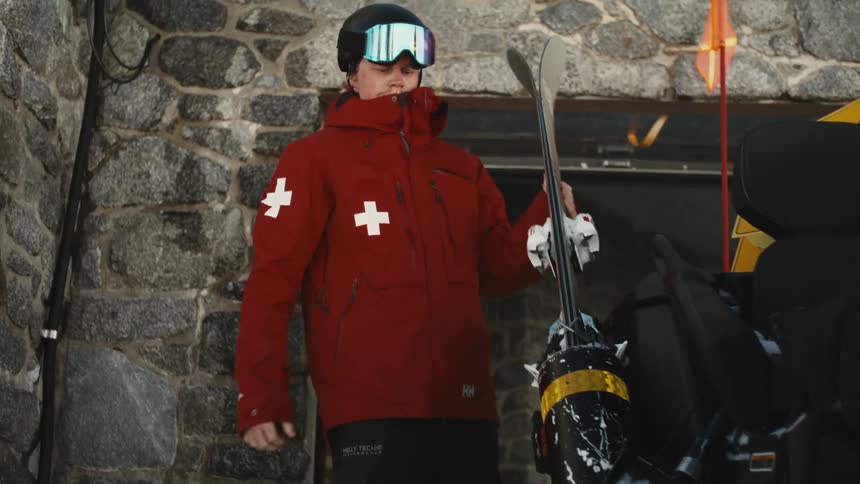 A ski patrol member in a red jacket holds their skis while standing next to a snowmobile outside a stone hut