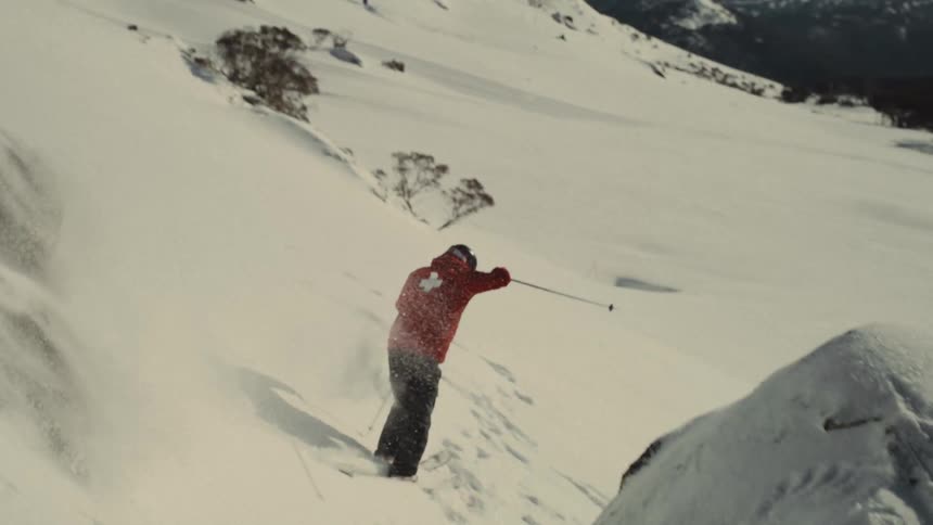 A skier in a red jacket carves through fresh powder down a snowy mountain in dramatic slow motion