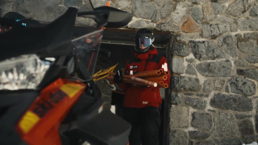 A member of the ski patrol carries safety equipment out of a stone hut towards a snowmobile in the winter