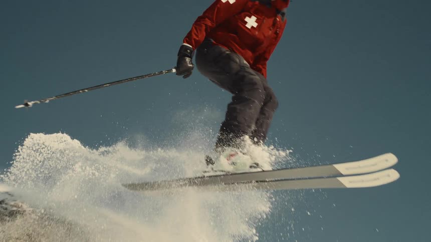 A professional skier in a red jacket and helmet jumps off a snowy rock in super slow motion, spraying powder snow into the air against a clear blue sky