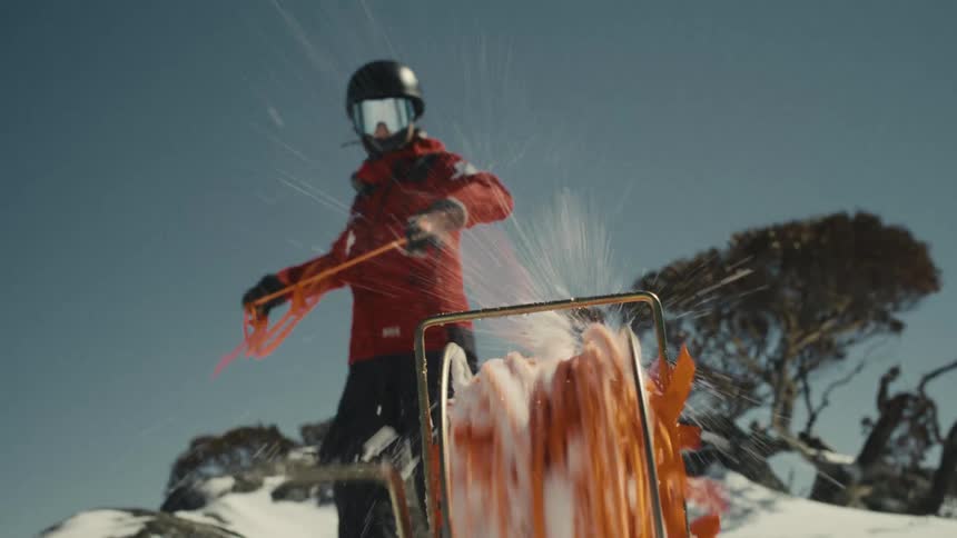 A mountain rescue worker in slow motion winds a bright orange safety line, spraying snow into the air on a sunny day