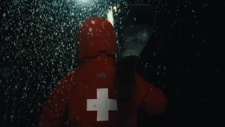 A person in a red hooded jacket with a white cross on the back carries skis over their shoulder in a heavy snowstorm at night