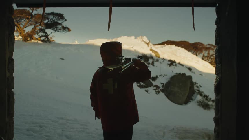 A silhouette of a ski patroller with skis on their shoulder looking out at a sunlit snowy mountain from inside a cabin
