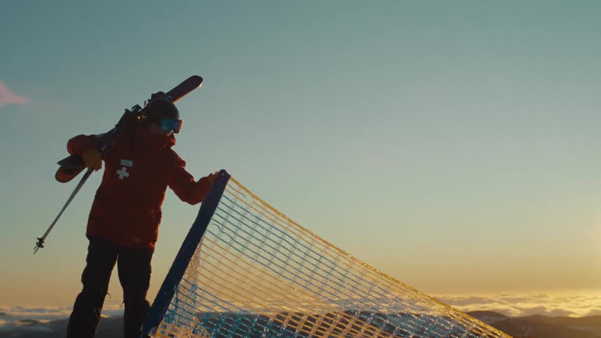 A ski patrol member carrying skis on their shoulder stands on a snowy mountain peak, overlooking a beautiful vista above the clouds at sunrise