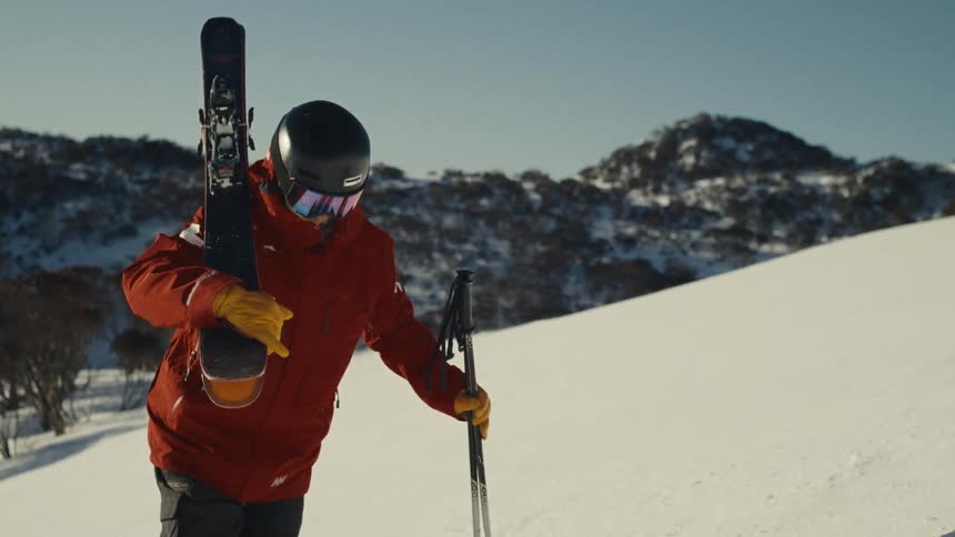 A skier in a red jacket and full gear carries his skis while standing on a sunny, snowy mountaintop